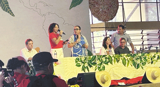 Image contains two alternating photos. The first
photo shows six people on stage, behind a table draped with a yellow tablecloth and decorated with woven hats. The second photo shows a large conference hall packed with people waving colorful flags.